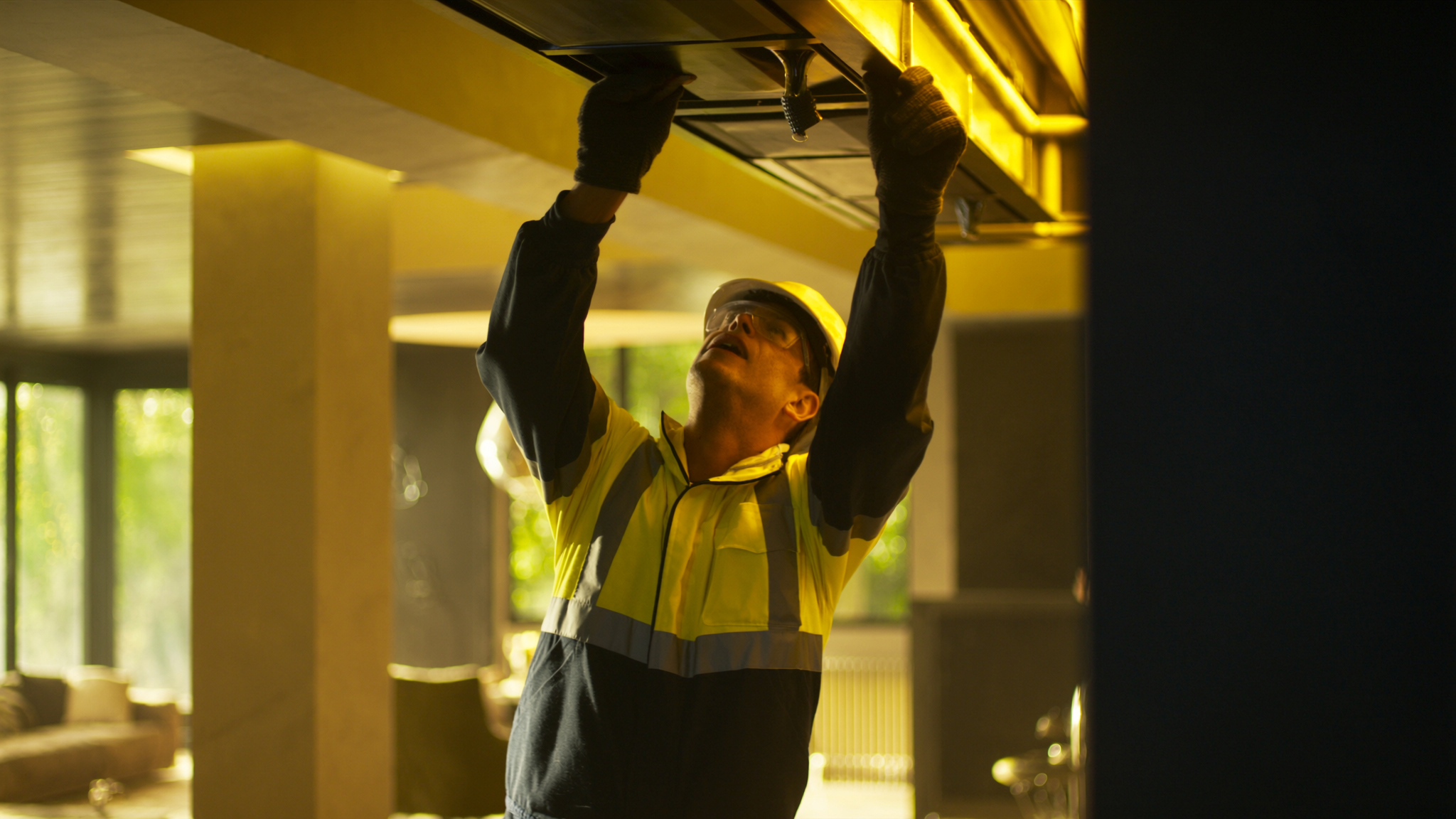 Worker in high-visibility vest installing ductwork on ceiling in modern interior space
