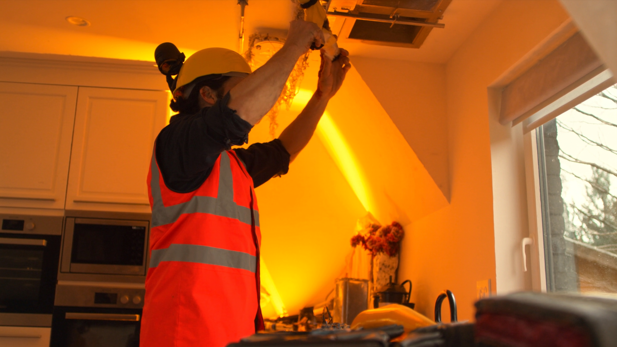 Worker in red safety vest installing ceiling fixture in orange-lit residential kitchen
