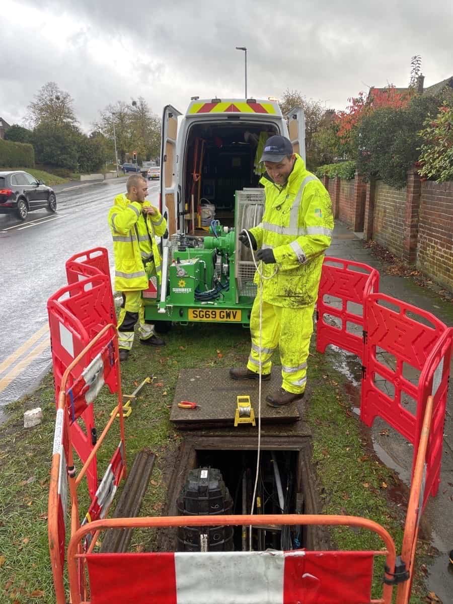 Two workers in high-visibility clothing at roadside access point with equipment and red barriers